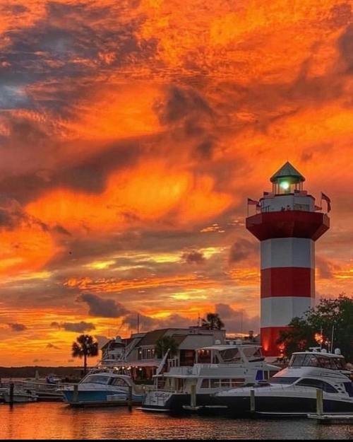 A vibrant sunset casts orange and pink hues over a striped lighthouse and boats in a marina.