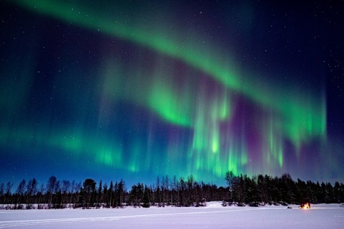 Vibrant auroras dance across a starry night sky over a snowy landscape, with a small cabin glowing in the distance.