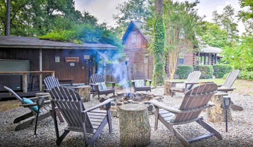 A cozy outdoor fire pit surrounded by wooden chairs, with a cabin and trees in the background. Smoke rises from the fire.