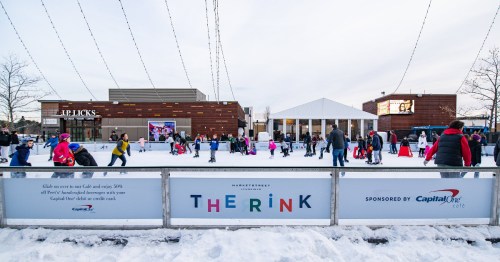 A busy ice skating rink with people skating and enjoying the winter atmosphere, featuring a large banner for Capital One.