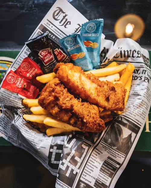 A plate of crispy fish fillets and golden fries, served with various dipping sauces on a newspaper backdrop.