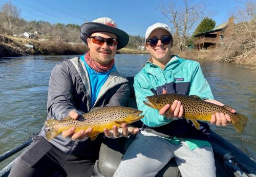 Two people in a boat hold up large brown trout, smiling against a sunny river backdrop.