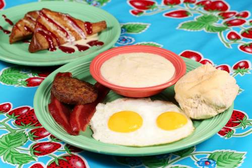A colorful plate featuring two fried eggs, sausage, grits, a biscuit, and sweet pastries drizzled with sauce.