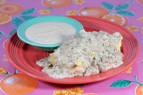 A plate of biscuits smothered in creamy sausage gravy, served with a side of grits, on a colorful floral tablecloth.
