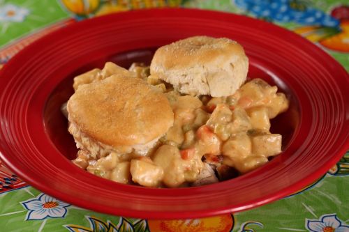 A red plate with two biscuits topped with creamy chicken and vegetable stew, set on a colorful patterned tablecloth.
