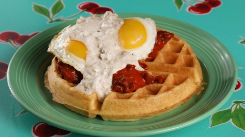 A waffle topped with fried chicken, creamy gravy, and two sunny-side-up eggs on a colorful cherry-patterned background.