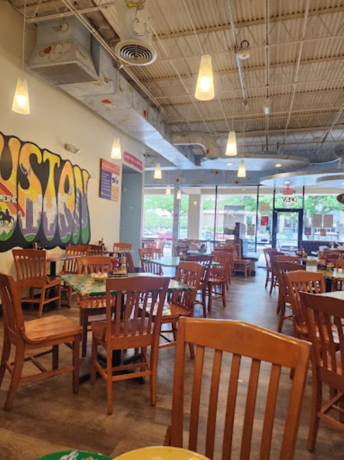 A spacious restaurant interior with wooden chairs and tables, colorful wall art, and large windows letting in natural light.