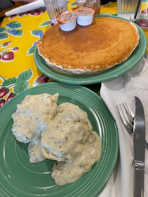 A plate with biscuits and gravy next to a large pancake topped with syrup cups, set on a colorful tablecloth.