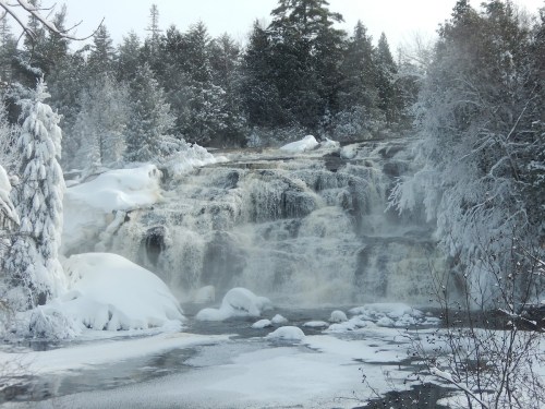 A snowy waterfall surrounded by frosted trees, cascading over rocks in a serene winter landscape.