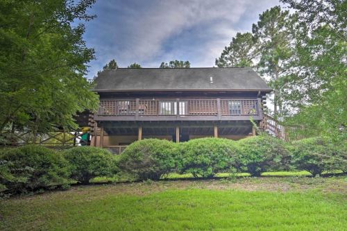 A two-story wooden house surrounded by greenery and shrubs, set against a backdrop of trees under a cloudy sky.
