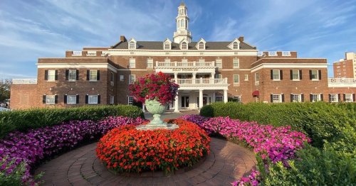 A grand brick building with a tower, surrounded by vibrant flower beds and a central fountain.
