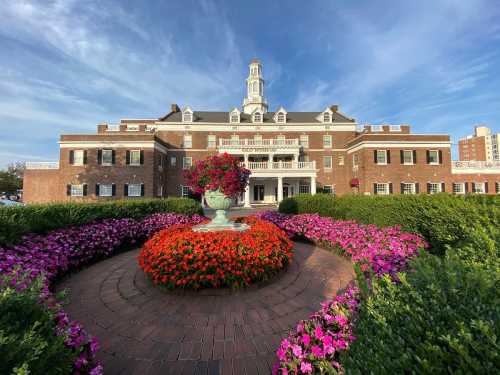 A grand brick building with a tower, surrounded by vibrant flower beds and lush greenery under a blue sky.