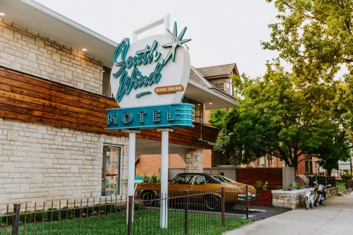 Sign for "South Wind Motel" with a vintage car parked outside, surrounded by greenery and a modern building.