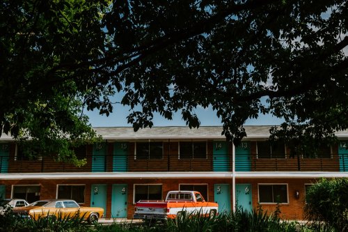 A vintage motel with turquoise doors and two classic cars parked outside, framed by leafy trees.