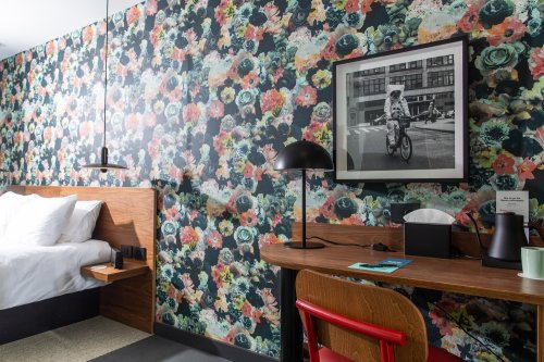 A stylish hotel room featuring floral wallpaper, a bed, a desk with a lamp, and a framed black-and-white photo.