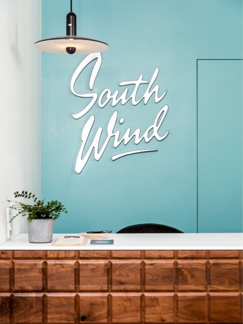 A modern reception area featuring a turquoise wall with "South Wind" signage and a wooden desk with a potted plant.