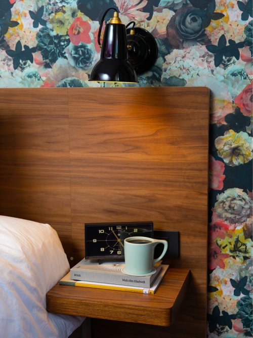 A bedside table with a clock, books, and a mug, against a floral wallpaper background. A lamp is mounted above.