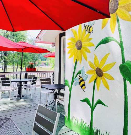 A patio with red umbrellas, tables, and a mural of sunflowers and bees on a white wall.