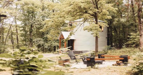 A round, metal-clad cabin surrounded by trees, with a fire pit and seating area in a serene forest setting.