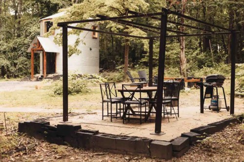 A rustic outdoor patio with a table and chairs, surrounded by trees, near a small building.
