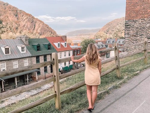 A woman in a yellow dress stands by a wooden fence, overlooking a scenic view of mountains and colorful houses.