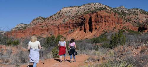 Three hikers walking on a trail with red rock formations and a clear blue sky in the background.