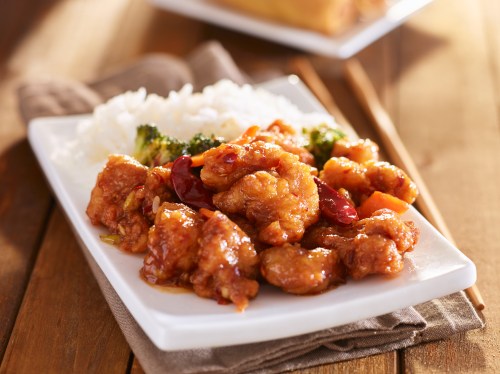 A plate of sweet and sour chicken with broccoli and white rice on a wooden table.