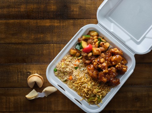 A takeout container with fried rice, noodles, sweet and sour chicken, and mixed vegetables, alongside a fortune cookie.