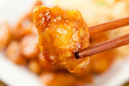 Close-up of a crispy, golden piece of chicken held with chopsticks, with a blurred background of fried rice.