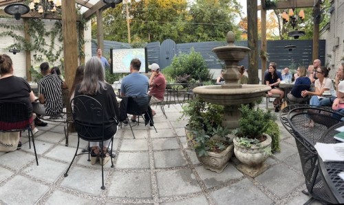 A group of people seated outdoors in a garden setting, listening to a speaker near a fountain.