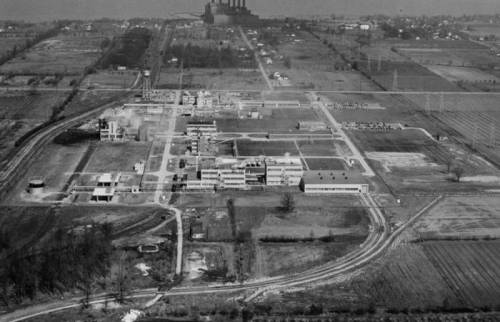 Aerial view of an industrial complex surrounded by fields and roads, with a power plant in the background.