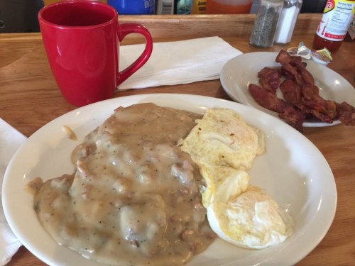 A plate of biscuits and gravy with two poached eggs, a side of bacon, and a red coffee mug on a wooden table.