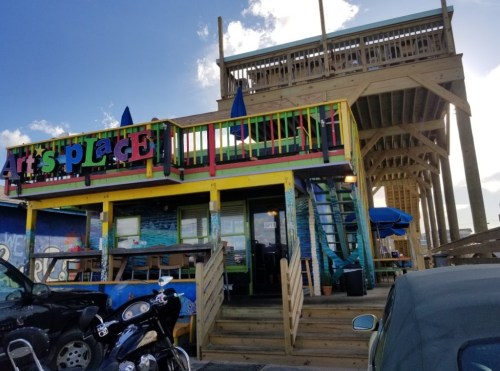 Colorful building with a wooden deck, outdoor seating, and a sign reading "Arts Place," surrounded by parked motorcycles.