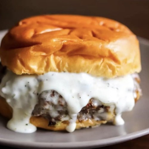A close-up of a juicy burger with creamy white sauce dripping from a soft, golden bun on a plate.