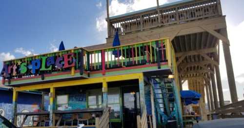 Colorful building with "Art's Place" sign, featuring outdoor seating and a deck above, under a blue sky.