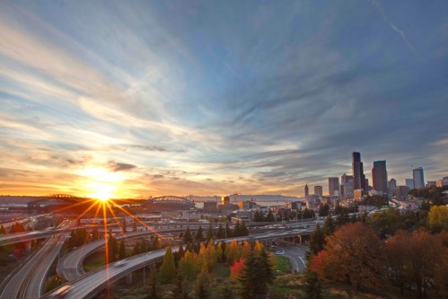 Sunset over a city skyline with highways and colorful autumn trees in the foreground.