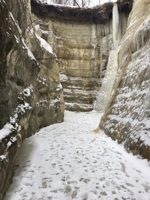 A snowy canyon with icy walls and a small waterfall, surrounded by steep, layered rock formations.