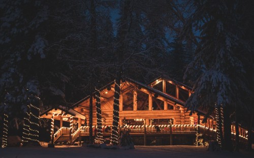 A cozy log cabin adorned with warm lights, surrounded by snow-covered trees at night.
