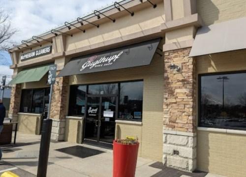Exterior of a storefront with a sign reading "Gyrothup" and a stone facade, featuring large windows and a red planter.