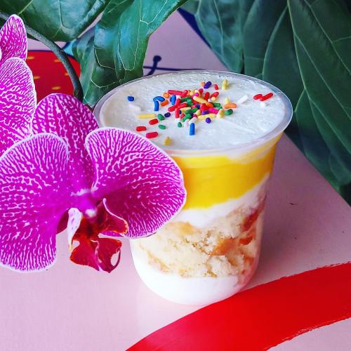 A colorful dessert in a cup with layers of cream, cake, and mango, topped with sprinkles and a purple orchid nearby.