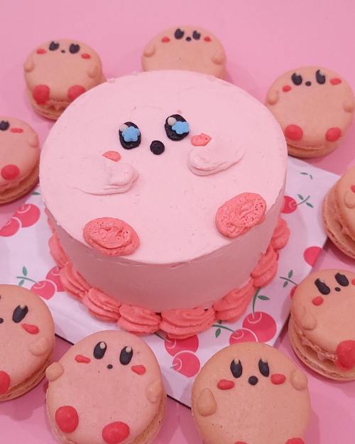 A pink cake decorated like Kirby, surrounded by Kirby-themed macarons on a pink background.