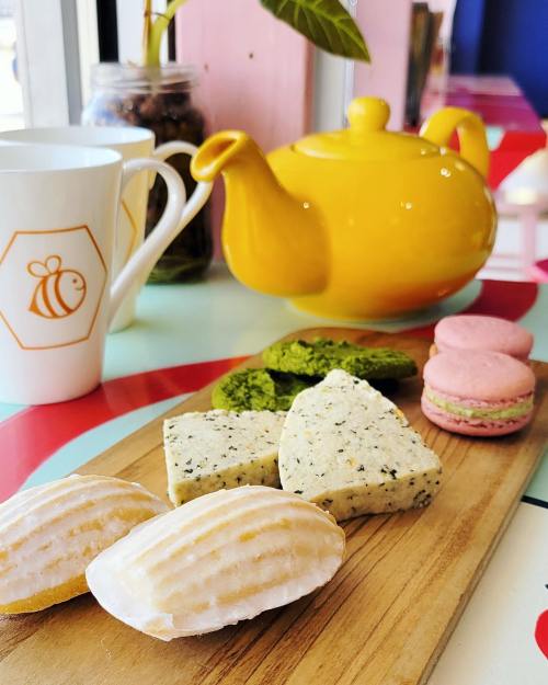 A wooden platter with madeleines, green tea sweets, and macarons, alongside a yellow teapot and a bee-themed mug.