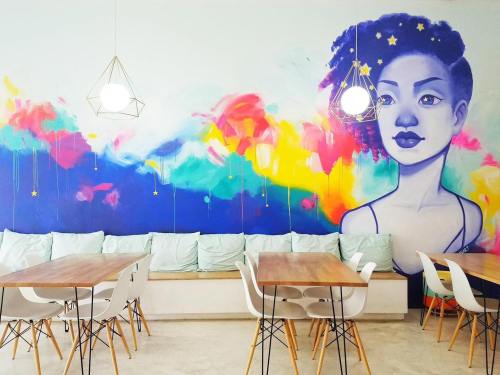 A vibrant mural of a woman with colorful abstract patterns, alongside modern tables and chairs in a cozy café setting.