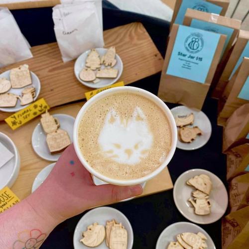 A hand holding a latte with a cat face latte art, surrounded by cookies and bags on a table.