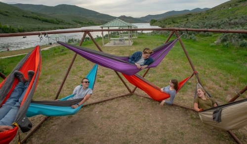 Four friends relax in colorful hammocks by a river, surrounded by green hills and a picnic shelter in the background.