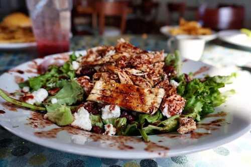 A colorful salad with shredded chicken, greens, goat cheese, and a balsamic dressing on a white plate.