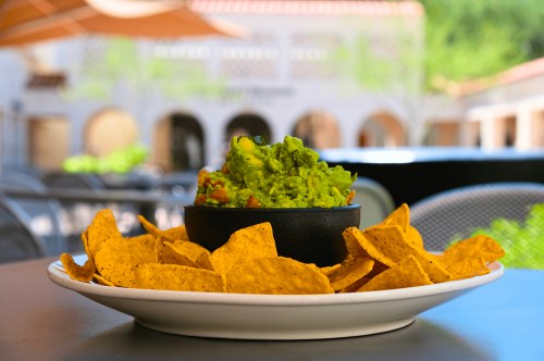 A plate of golden tortilla chips surrounds a bowl of fresh guacamole, set outdoors with a blurred background.