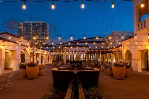 A beautifully lit courtyard at dusk, featuring string lights, potted plants, and a central fountain.