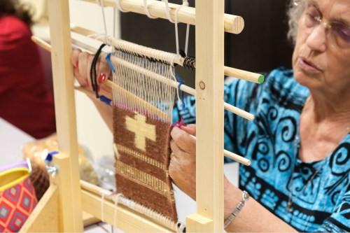 An elderly woman weaves on a loom, creating a textile with a cross design, focused on her craft.
