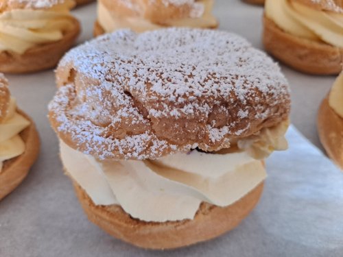 A close-up of a cream-filled pastry topped with powdered sugar, surrounded by similar pastries on a tray.